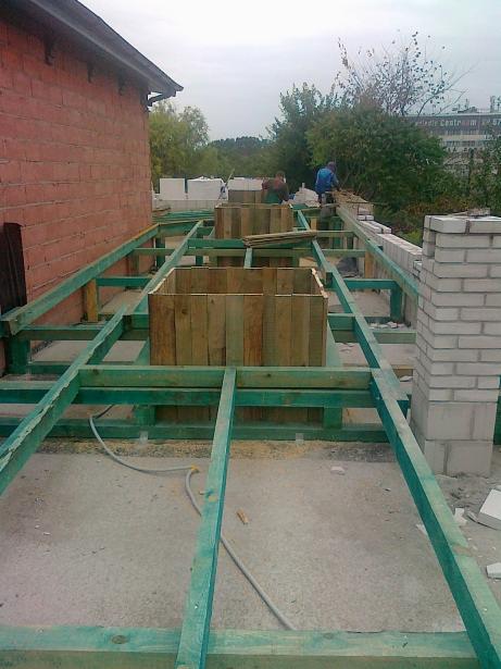 Wiązanie zbrojenia pod fundament z drewnianymi szalunkami na budowie domu. W tle mur z cegły i pracujący robotnicy.