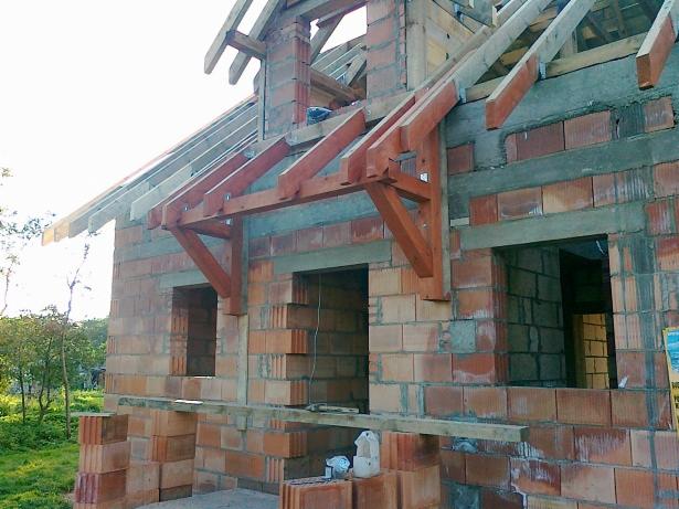 Surowy stan domu z czerwonej cegły z drewnianą konstrukcją dachu i nadprożami okiennymi, w trakcie budowy, widok z zewnątrz w słoneczny dzień.