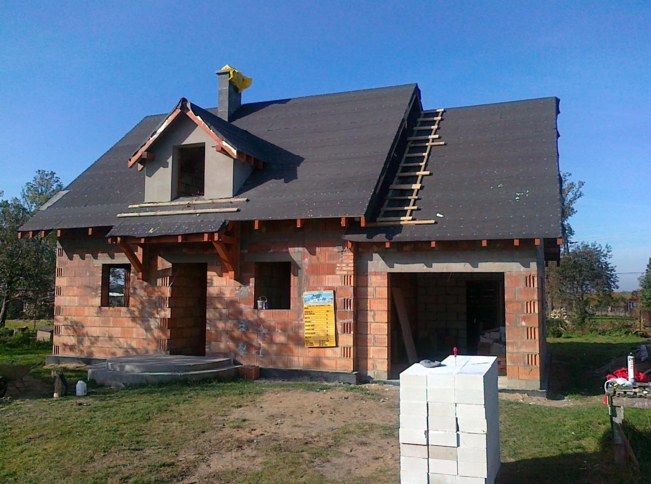 Niedokończony dom murowany z czerwonej cegły z czarnym dachem. Widoczne pustaki, okna bez szyb, drabina na dachu. Słoneczny dzień.