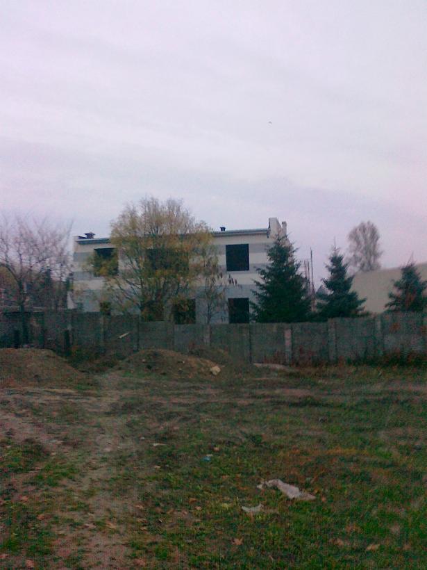 Niedokończony budynek dwukondygnacyjny z widoczną surową elewacją i czarnymi otworami okiennymi, otoczony ogrodzeniem i drzewami w pochmurny dzień.