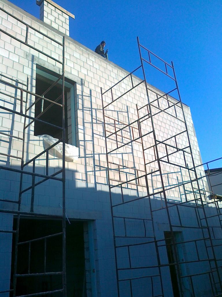 Budowa domu z białej cegły silikatowej, widoczne rusztowania i pracownik na dachu. Stan surowy otwarty, okno bez ramy.