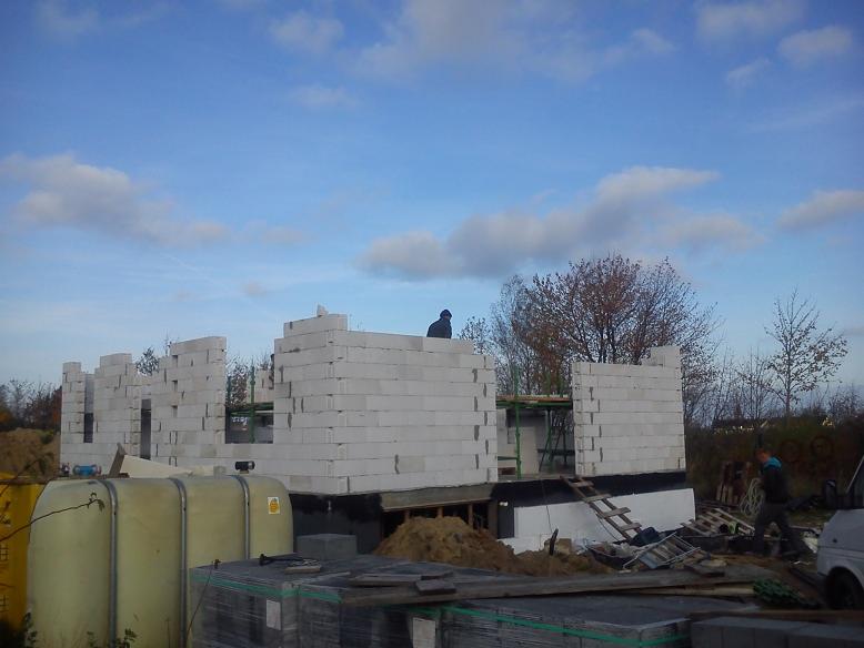 Budowa domu w trakcie: wznoszone ściany z bloczków, widoczny pracownik na rusztowaniu i materiały budowlane. Słoneczny dzień, błękitne niebo z chmurami. Jesienne otoczenie.