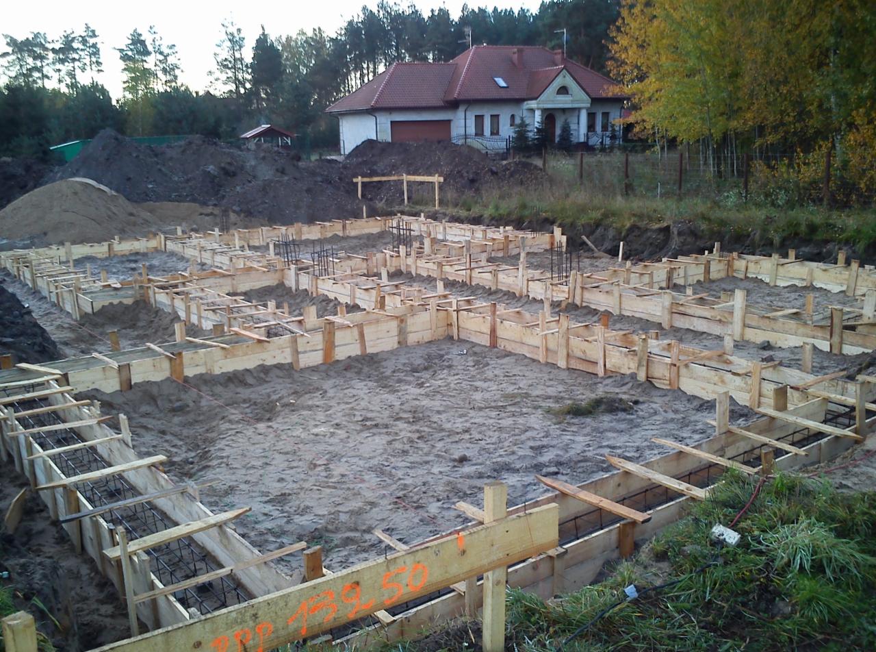 Szczegółowe deskowanie pod fundamenty domu jednorodzinnego, widoczne zbrojenie, w tle budynek w stanie surowym otwartym i hałda ziemi.