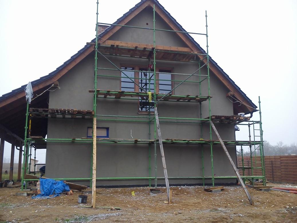 Dom w trakcie tynkowania z zielonym rusztowaniem. Widoczna szara elewacja, okna i fragment dachu. Na ziemi porozrzucane materiały budowlane. Pochmurna pogoda.