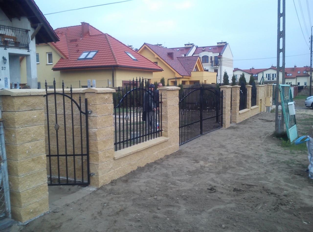Nowe ogrodzenie z kamiennymi słupkami i metalową bramą, widok na posesje z domami w tle. Elementy dekoracyjne na bramie. Teren nieutwardzony.