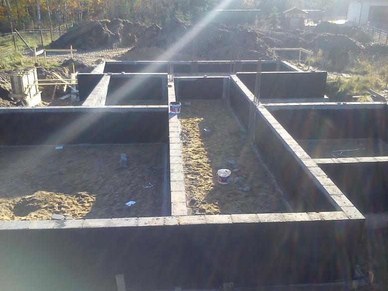 Zalane fundamenty domu z bloczków betonowych, widoczne zbrojenia i piasek wewnątrz. W tle ziemia i drzewa, słoneczny dzień.
