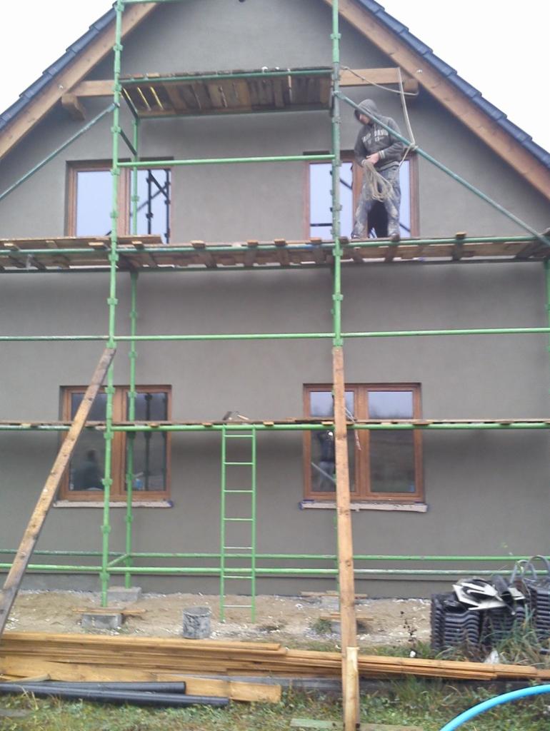 Prace tynkarskie na elewacji domu jednorodzinnego z użyciem rusztowania. Widoczny pracownik na rusztowaniu z liną. Tynk w kolorze szarym.