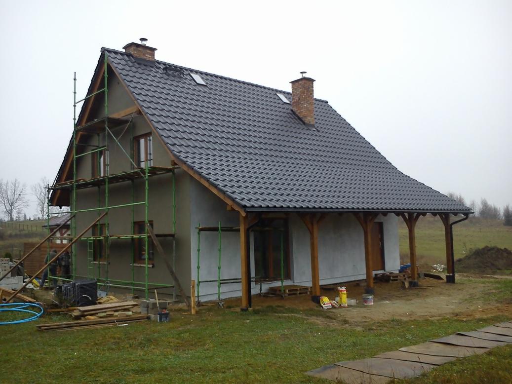 Dom jednorodzinny w trakcie prac elewacyjnych, z widocznymi rusztowaniami i świeżo otynkowaną ścianą. Dach pokryty ciemną dachówką.