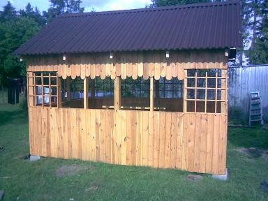 Drewniana altana ogrodowa z brązowym dachem i dekoracyjnymi wykończeniami. Widoczne okna z kratkami, zielona trawa i drzewa w tle. Konstrukcja na betonowych bloczkach.