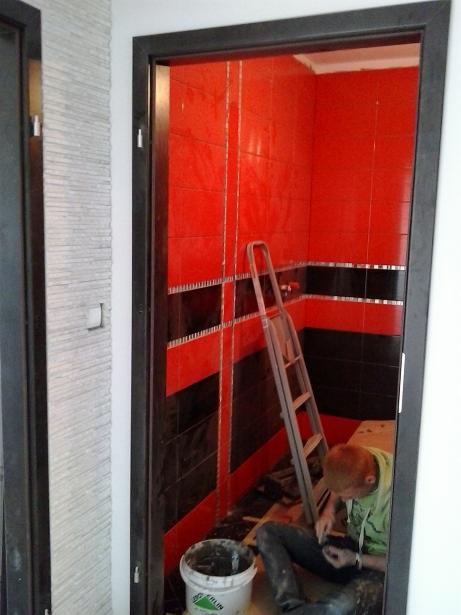 Układanie czerwono-czarnych płytek w łazience. Widoczny glazurnik przy pracy, drabina oparta o ścianę i wiadro z zaprawą. Nietypowe połączenie kolorów.