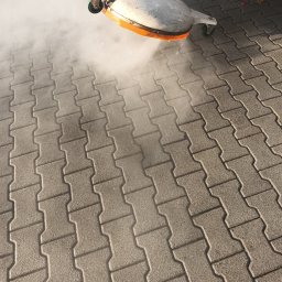 Usługi Wysokościowe FHU Mix Bud Piotr Smagacz - Czyszczenie kostki brukowej za pomocą myjki ciśnieniowej z okrągłą dyszą, widoczna para wodna i czyszczona powierzchnia.