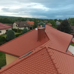 Usługi Wysokościowe FHU Mix Bud Piotr Smagacz - Czerwony dach pokryty dachówką bitumiczną w kształcie rybiej łuski, z widocznym kominem z cegły i oknem dachowym, na tle zieleni i zabudowań pod pochmurnym niebem.