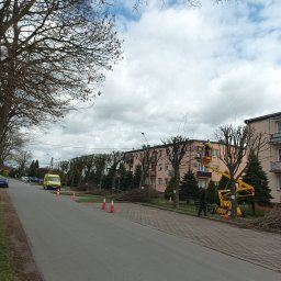 Usługi Wysokościowe FHU Mix Bud Piotr Smagacz - Pracownik w żółtym kombinezonie przycina drzewa z podnośnika na tle bloku mieszkalnego, z gałęziami leżącymi na chodniku i ulicy, zabezpieczonymi pomarańczowymi pachołkami.