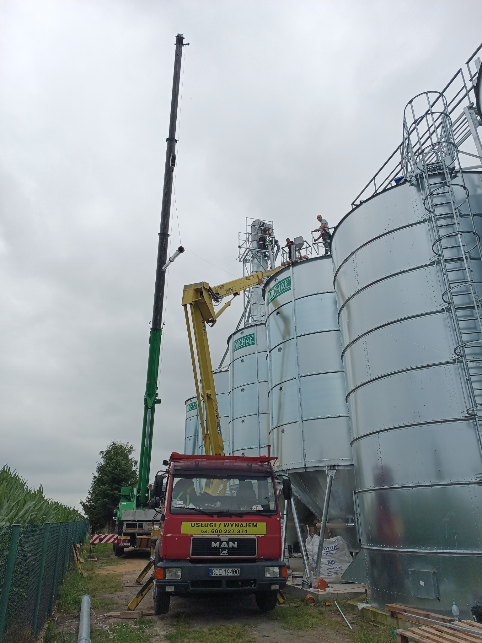 Montaż silosów zbożowych z użyciem dźwigu, trzech pracowników na szczycie konstrukcji, widoczna ciężarówka MAN z żółtą naklejką informacyjną.
