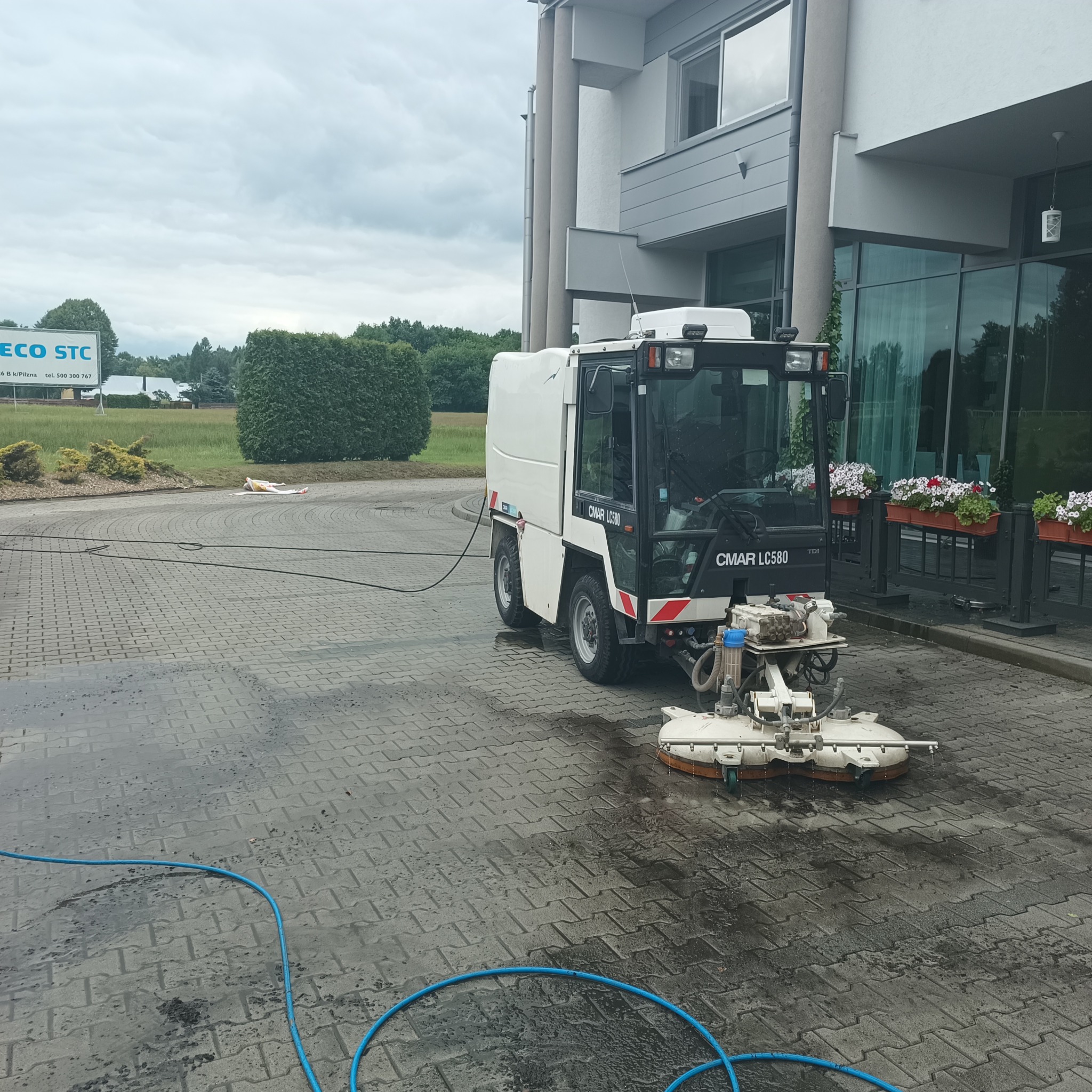 Maszyna CMAR LC580 w trakcie czyszczenia mokrej kostki brukowej przed budynkiem, widoczny niebieski wąż doprowadzający wodę.