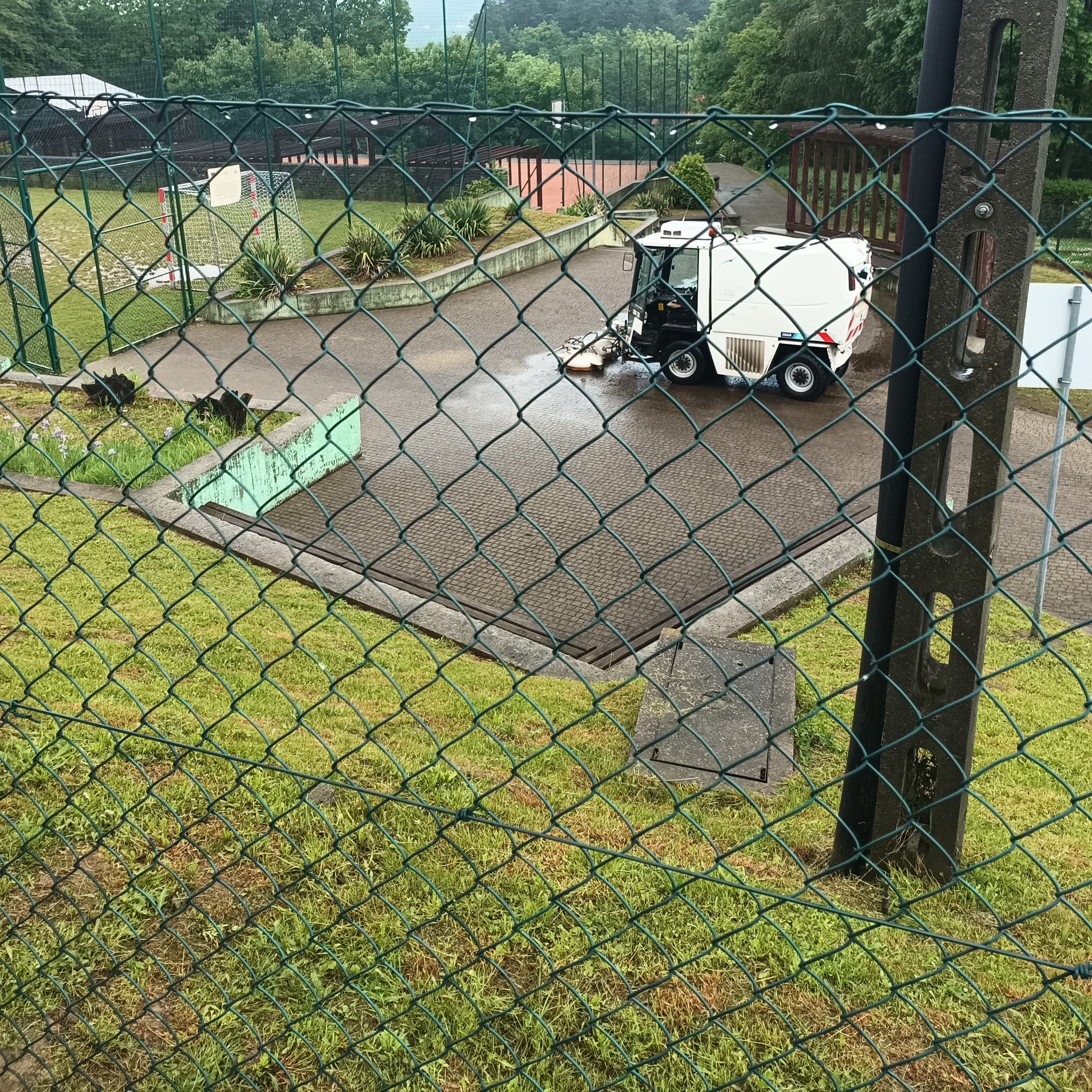 Maszyna czyszcząca kostkę brukową na mokro, widoczna przez siatkę ogrodzeniową na tle boiska sportowego.