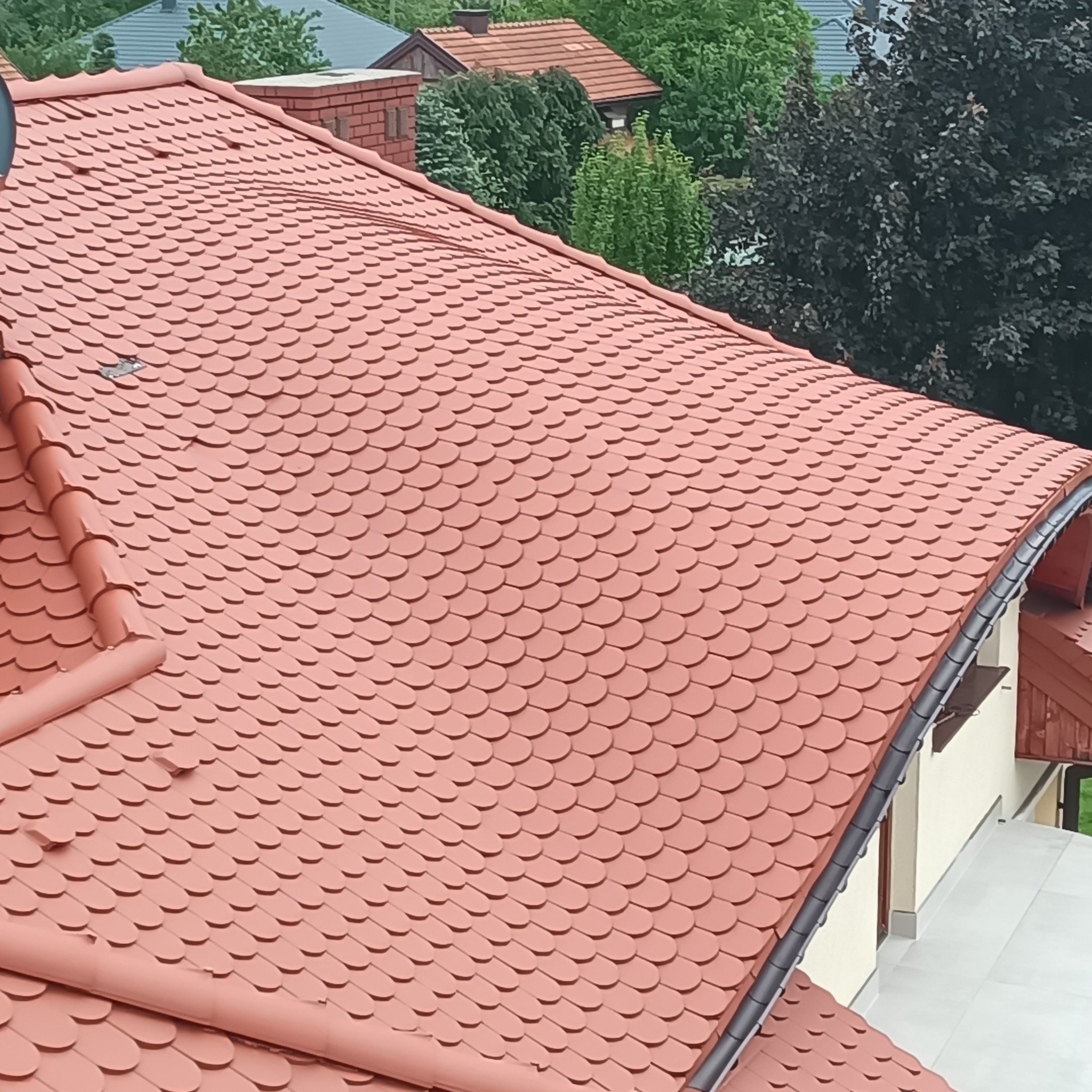 Dach pokryty czerwoną dachówką karpiówką, widok z góry, zarys komina i zieleni w tle.