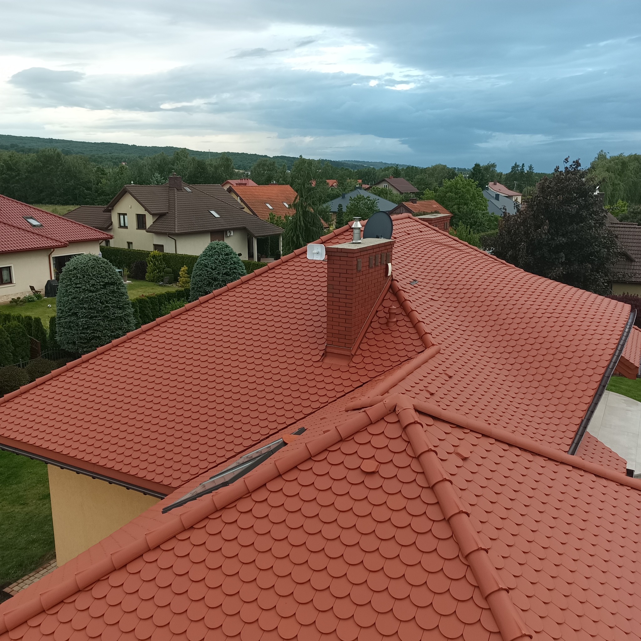 Czerwony dach pokryty dachówką bitumiczną w kształcie rybiej łuski, z widocznym kominem z cegły i oknem dachowym, na tle zieleni i zabudowań pod pochmurnym niebem.
