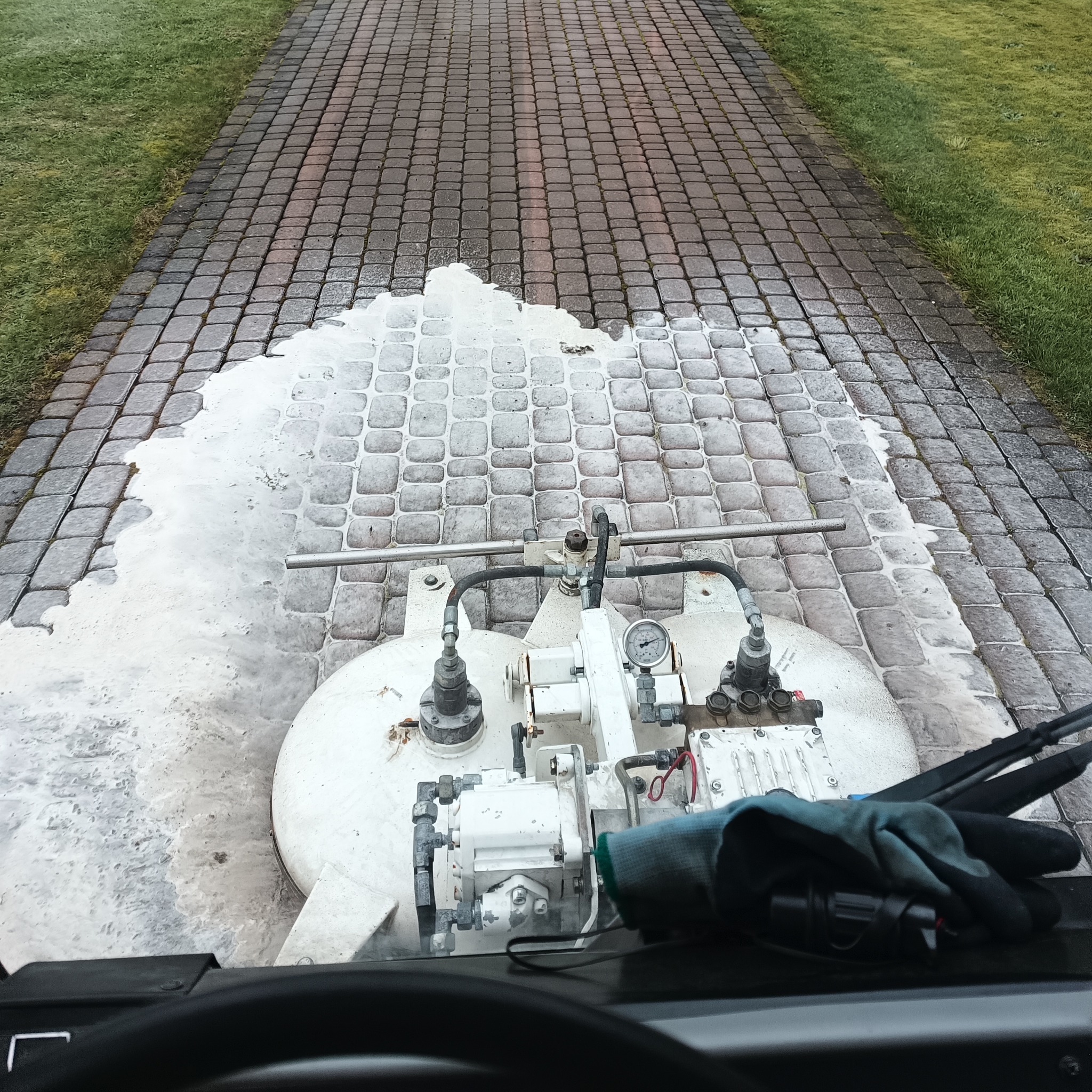 Maszyna czyszcząca kostkę brukową w trakcie pracy, widok z perspektywy operatora, część kostki już umyta, w tle trawnik.