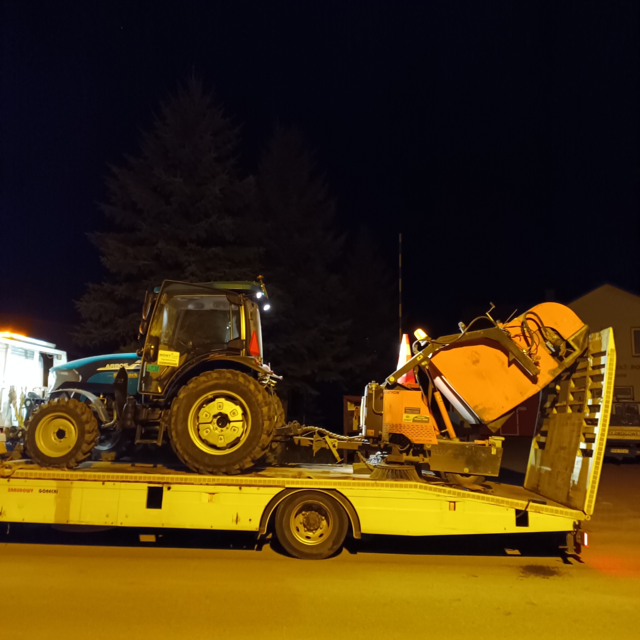 Ciężarówka z platformą transportową w Starej Jastrząbce przewozi traktor z zamontowaną szczotką do zamiatania, prawdopodobnie do prac porządkowych.