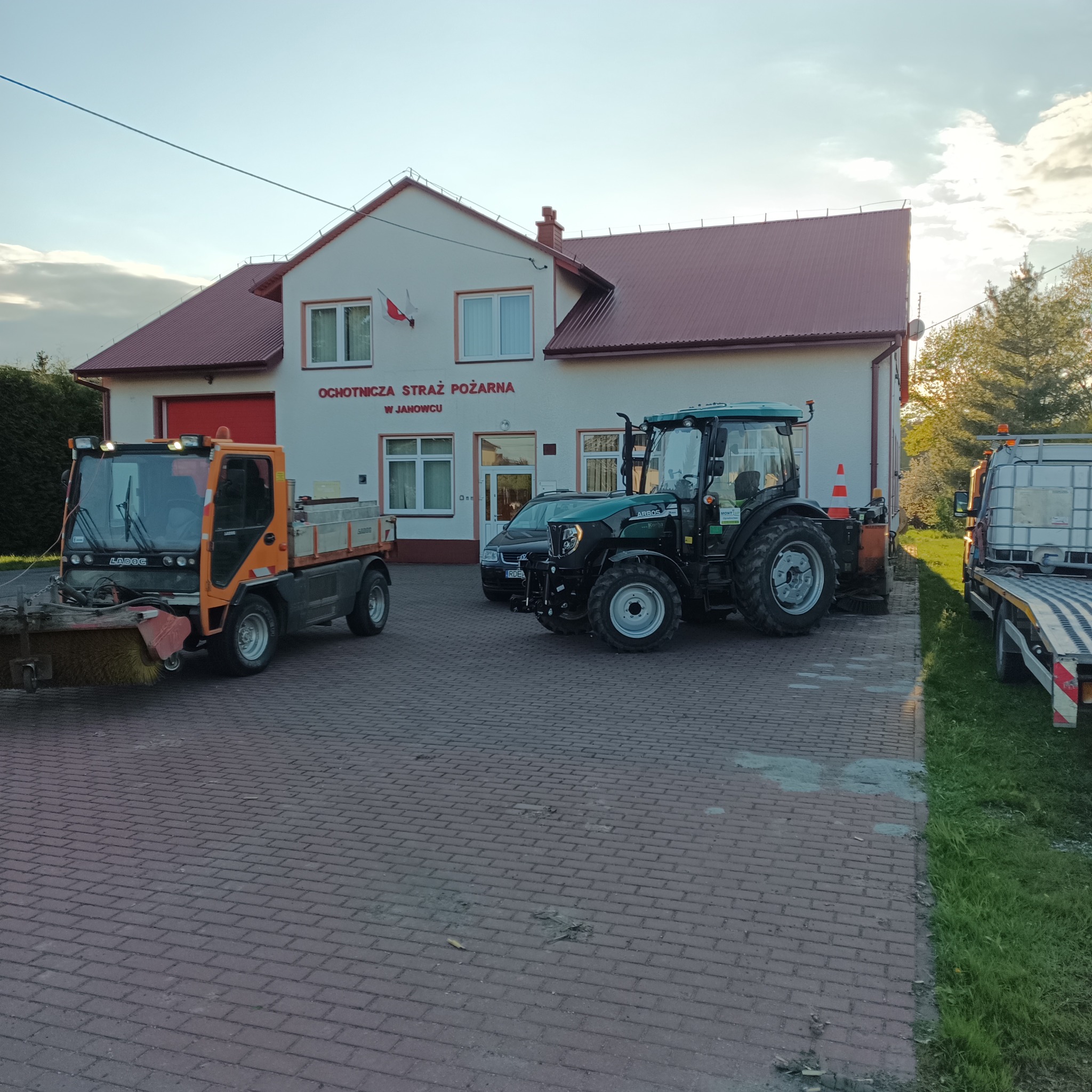 Pomarańczowa zamiatarka uliczna i zielony traktor z szczotką do odśnieżania przed budynkiem Ochotniczej Straży Pożarnej w Janowcu, kostka brukowa na pierwszym planie.