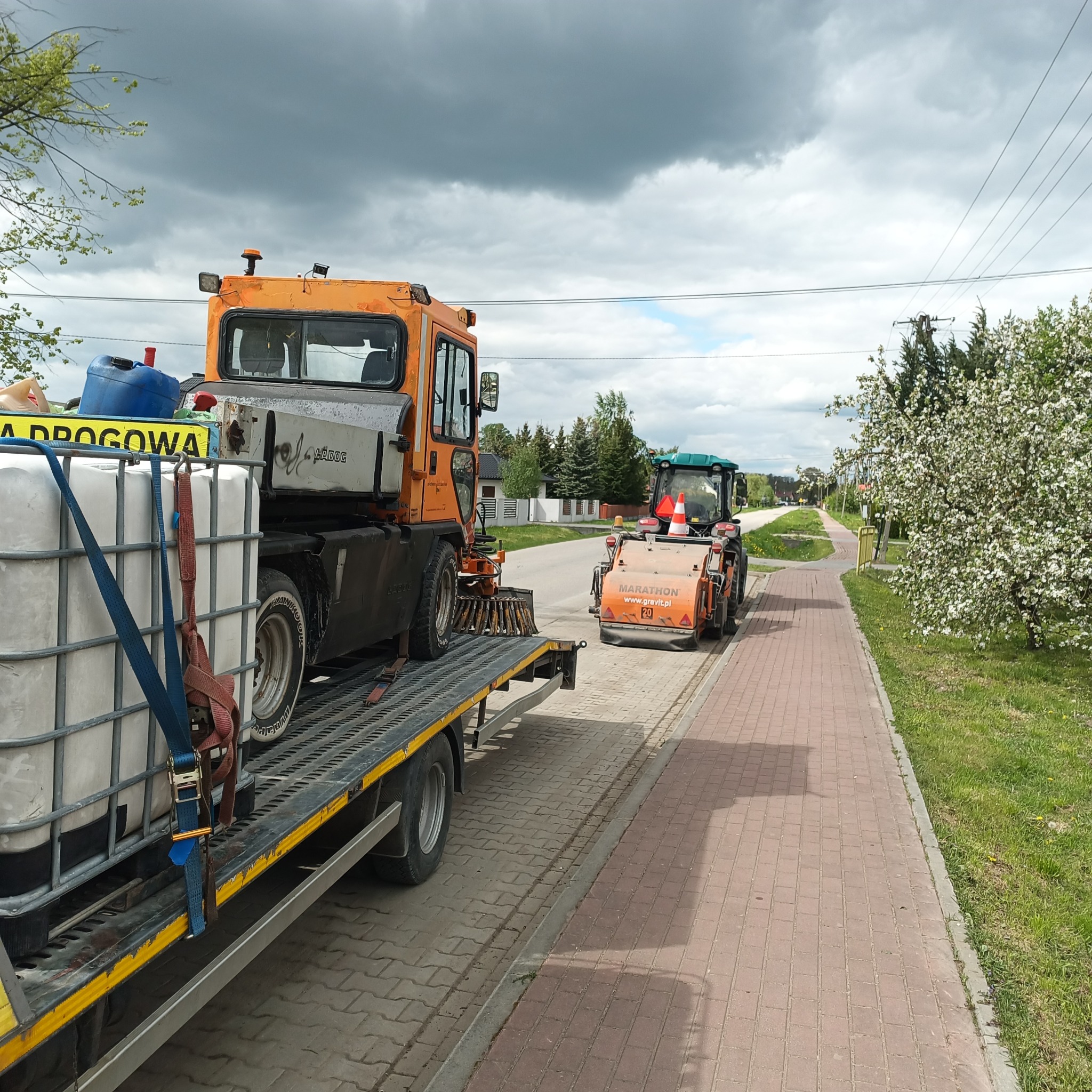 Pomarańczowy pojazd do czyszczenia dróg z szczotkami i zbiornikiem na platformie, za nim walec drogowy na chodniku z kostki brukowej, w tle kwitnące drzewo i pochmurne niebo.