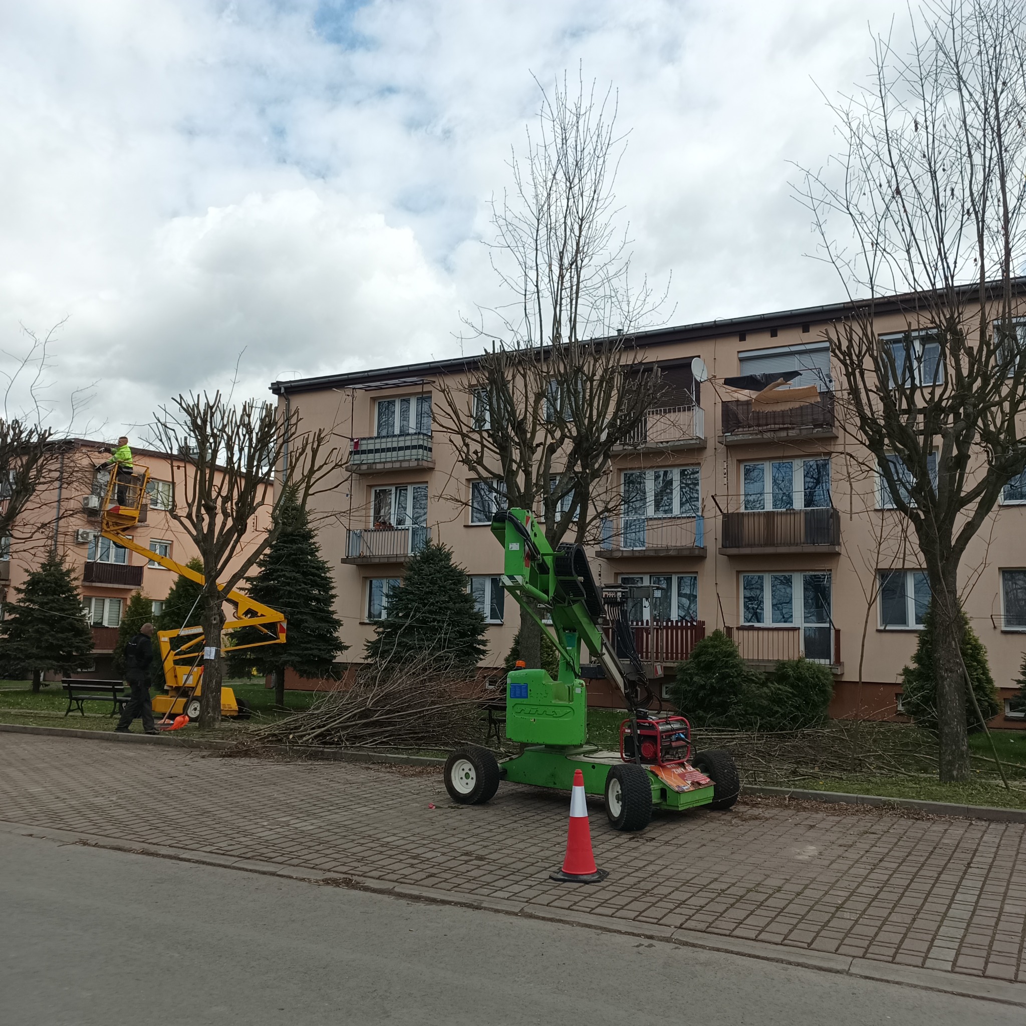 Pracownicy w żółtych podnośnikach koszowych przycinają gałęzie drzew przed blokiem mieszkalnym, obok leżą ścięte gałęzie, a zielona maszyna rozdrabnia odpady.