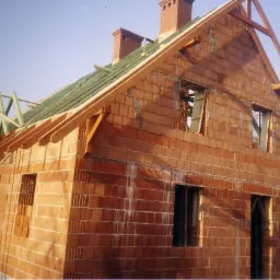 Budowa domu jednorodzinnego w trakcie wznoszenia ścian z czerwonej cegły, widoczna konstrukcja dachu i kominy. Ujęcie z dołu, w tle błękitne niebo i drzewa.