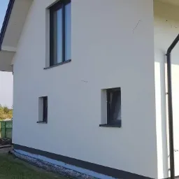 Jasna elewacja domu jednorodzinnego z grafitowymi oknami i dachem, widoczne ślady po niedawnym tynkowaniu na ścianie.