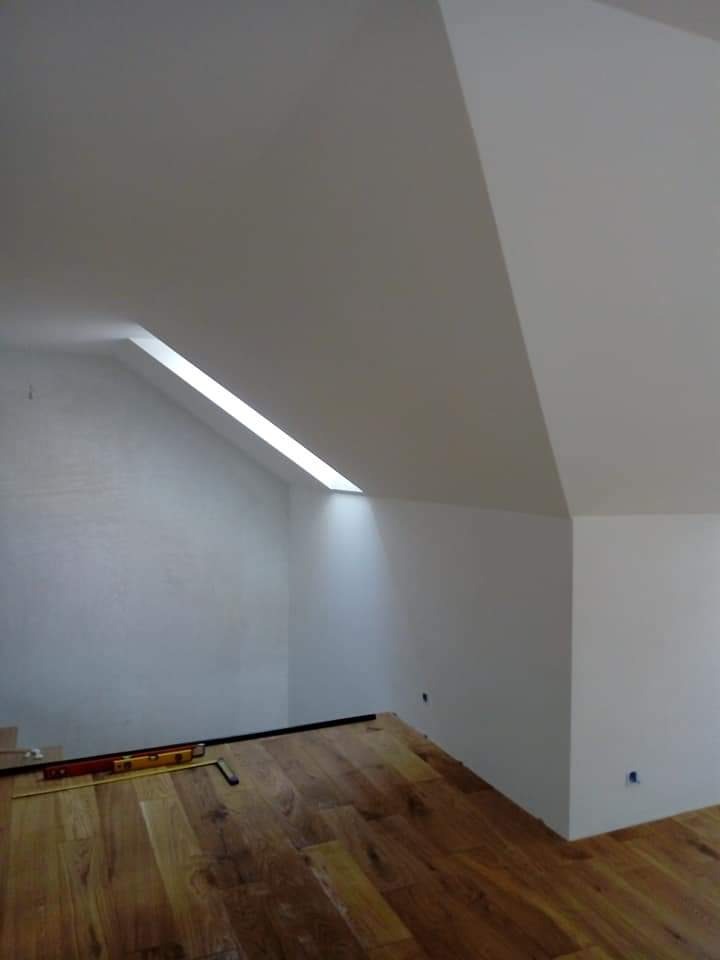Poddasze z nową, białą ścianą i drewnianą podłogą; poziomica i miara na podłodze sugerują prace wykończeniowe.