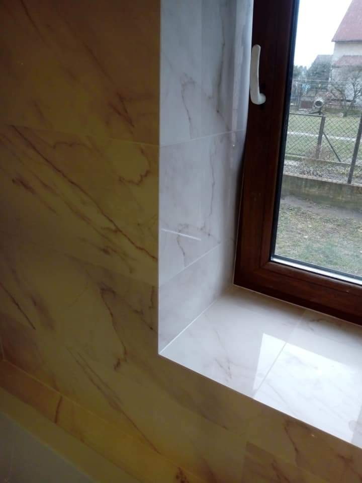 Wykończone wnęki okienne wewnątrz budynku, obłożone płytkami imitującymi marmur w odcieniach beżu i bieli, z widocznym brązowym oknem PCV i widokiem na ogród.