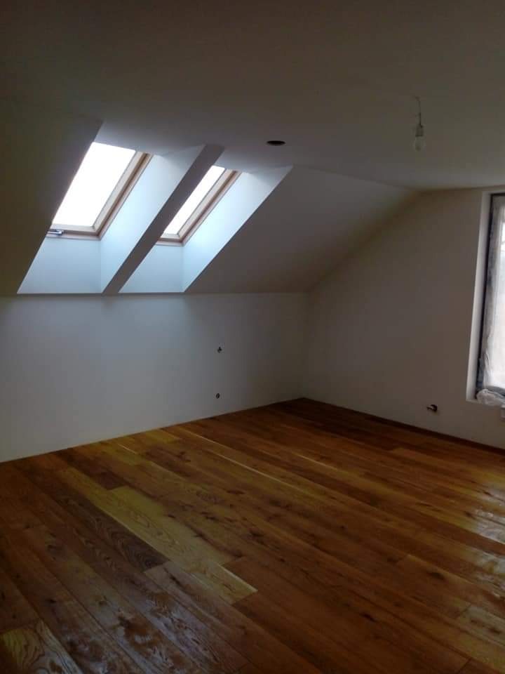 Poddasze z drewnianą podłogą i dwoma oknami dachowymi, świeżo pomalowane ściany w odcieniach bieli, widoczne niedoskonałości malowania przy oknach.