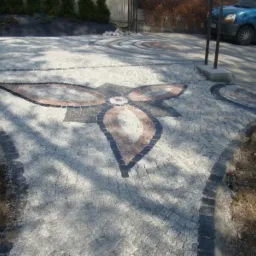 Nawierzchnia z jasnoszarej kostki z dekoracyjnym wzorem kwiatowym z ciemniejszej kostki, widoczny fragment samochodu w tle.