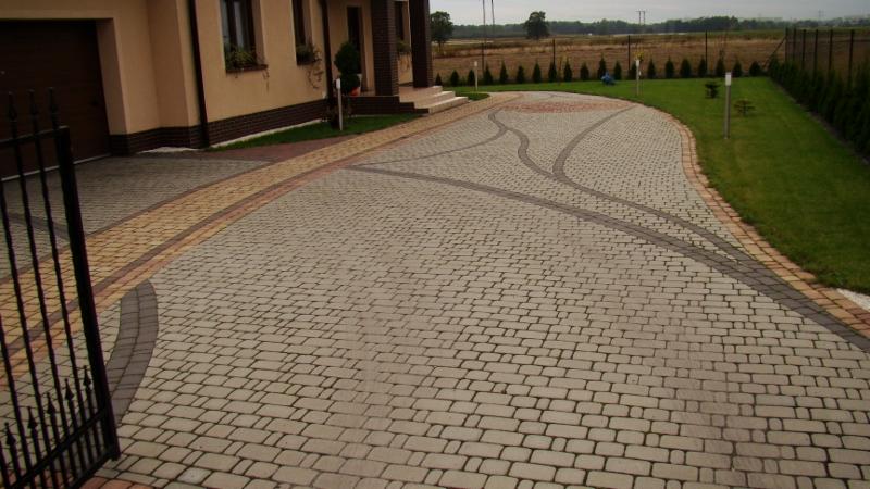 Szeroki podjazd przed domem wyłożony beżową kostką brukową z dekoracyjnymi wstawkami z ciemniejszej kostki, otoczony zielonym trawnikiem i ogrodzeniem.