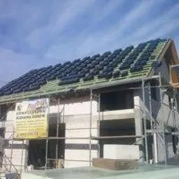 Budowa domu w trakcie realizacji: wzniesione ściany z pustaków, częściowo pokryty dach z czarną dachówką, widoczne rusztowania i baner reklamowy firmy budowlanej.