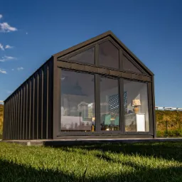 Nowoczesny, czarny budynek z dużymi przeszkleniami i dwuspadowym dachem, na trawniku, z widokiem na błękitne niebo i skarpę z zielenią w tle.