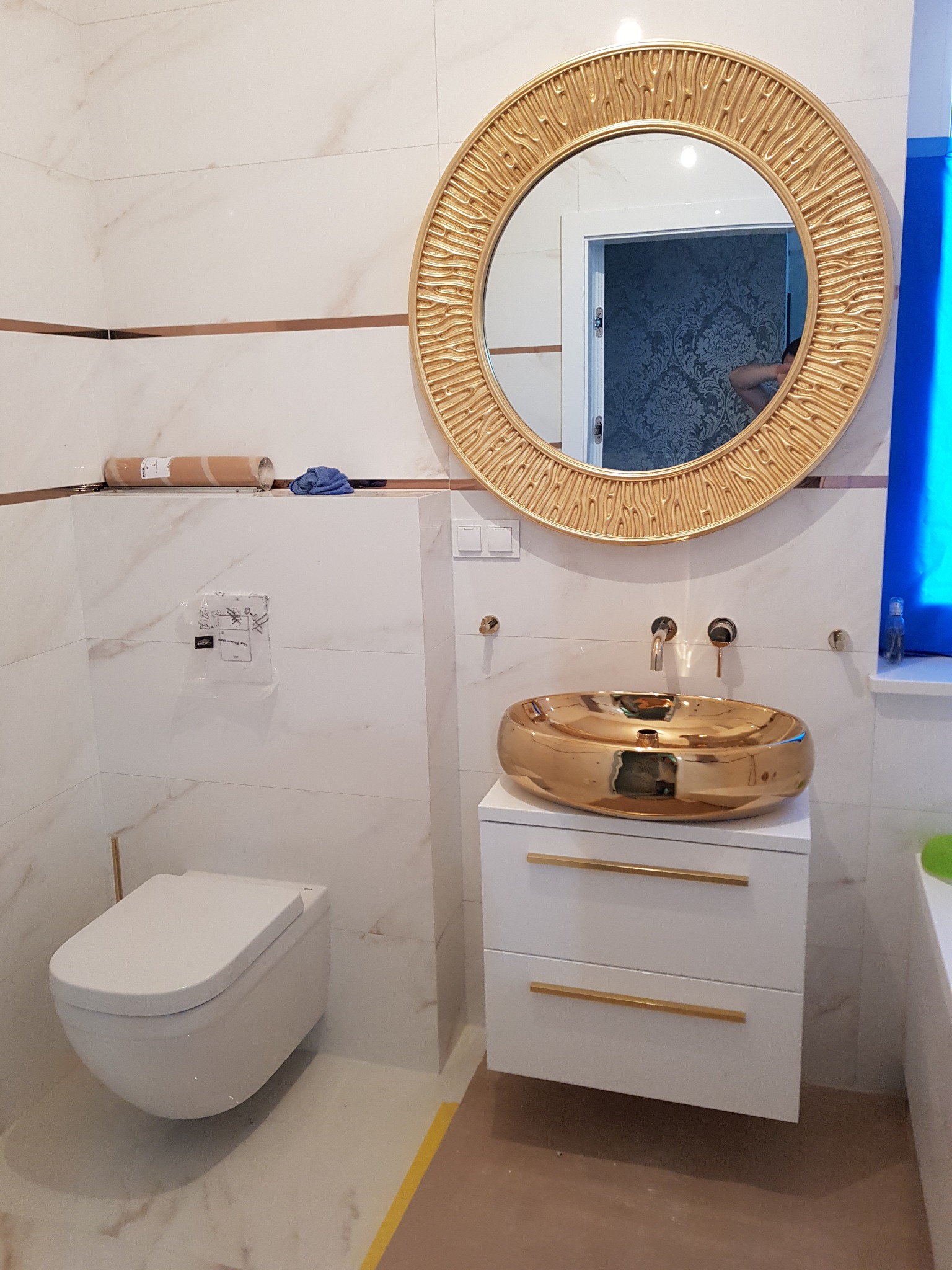 Nowoczesna łazienka z marmurowymi płytkami, złotą okrągłą umywalką nablatową i lustrem z dekoracyjną ramą w kolorze złota, podwieszana toaleta.