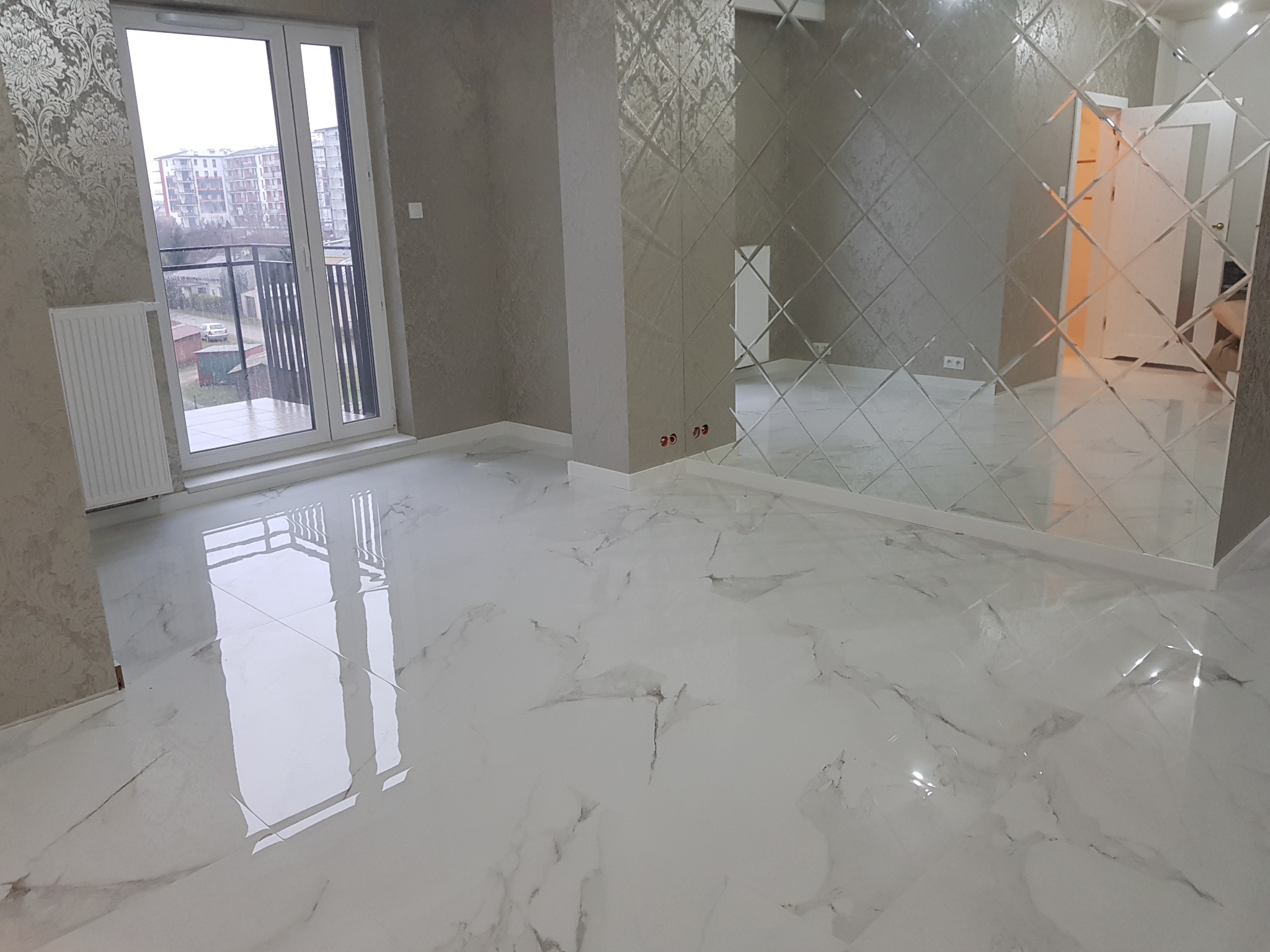 Wykończone wnętrze pokoju z błyszczącą, marmurkową podłogą, ścianą z lustrzanych rombów i balkonem z widokiem na budynki mieszkalne.