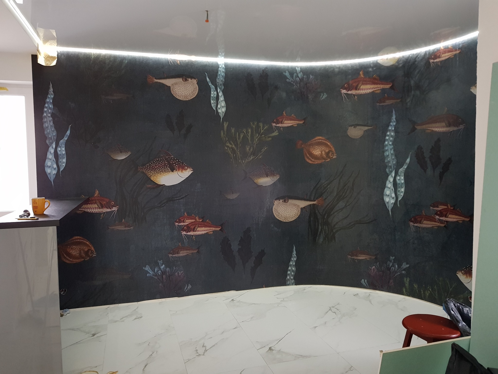 Pokój po remoncie z tapetą w motywy morskie, oświetleniem LED pod sufitem i marmurową podłogą. Widoczny fragment mebla z kubkiem i małym przedmiotem na blacie, oraz czerwony stołek w rogu.