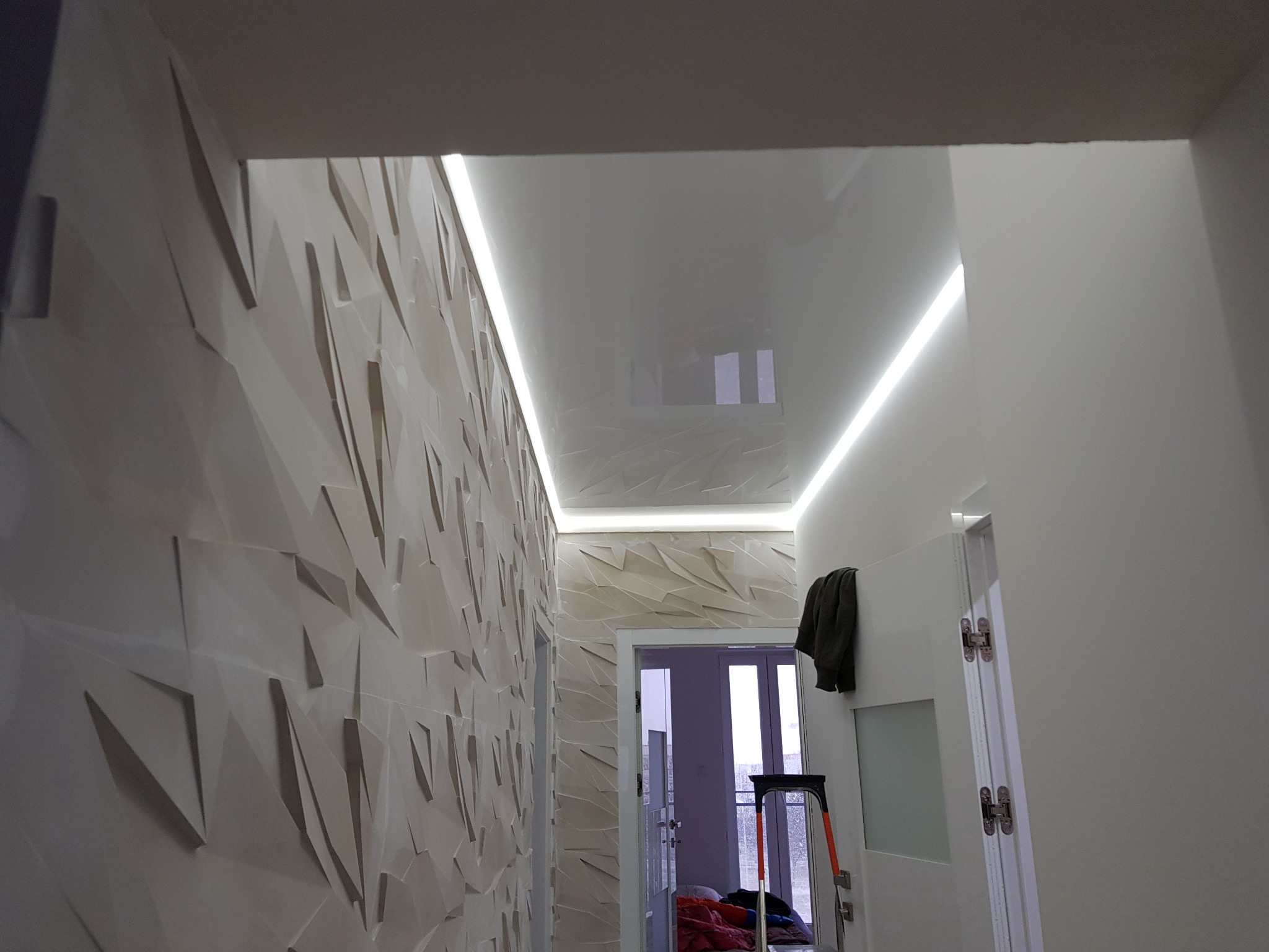 Nowoczesny korytarz z trójwymiarową ścianą w odcieniach beżu, podświetlonym sufitem i widoczną drabiną malarską w tle, sugerującą prace wykończeniowe.