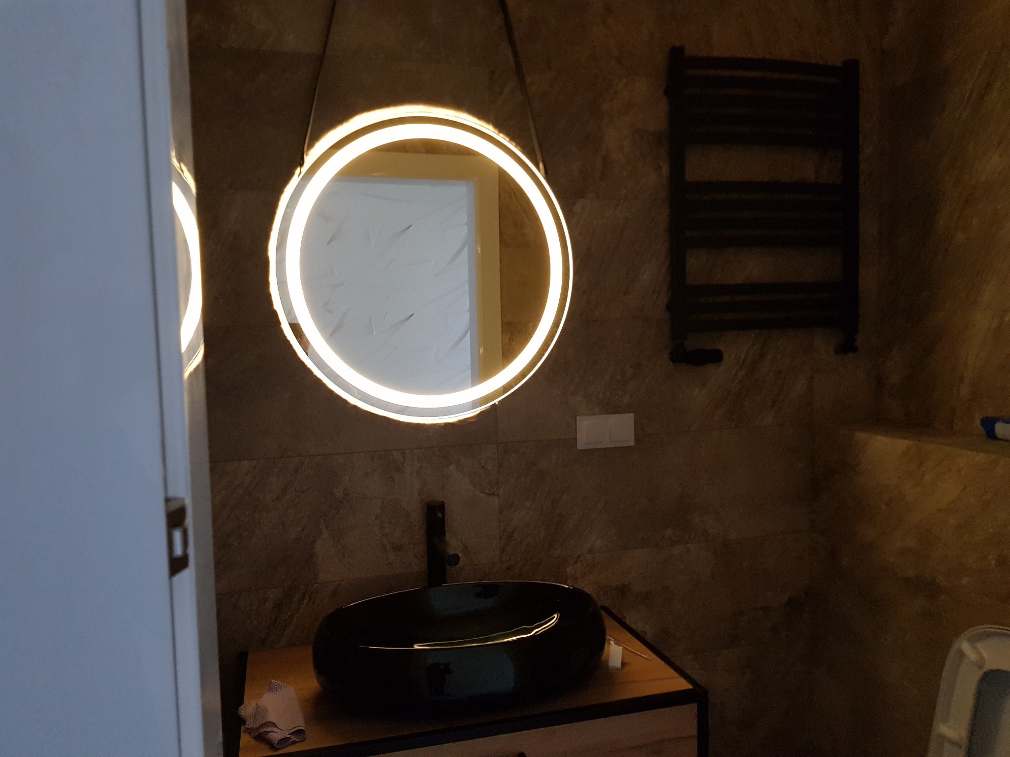 Łazienka z okrągłym lustrem podświetlanym LED, czarną umywalką nablatową i grzejnikiem drabinkowym na ścianie z imitacją kamienia.