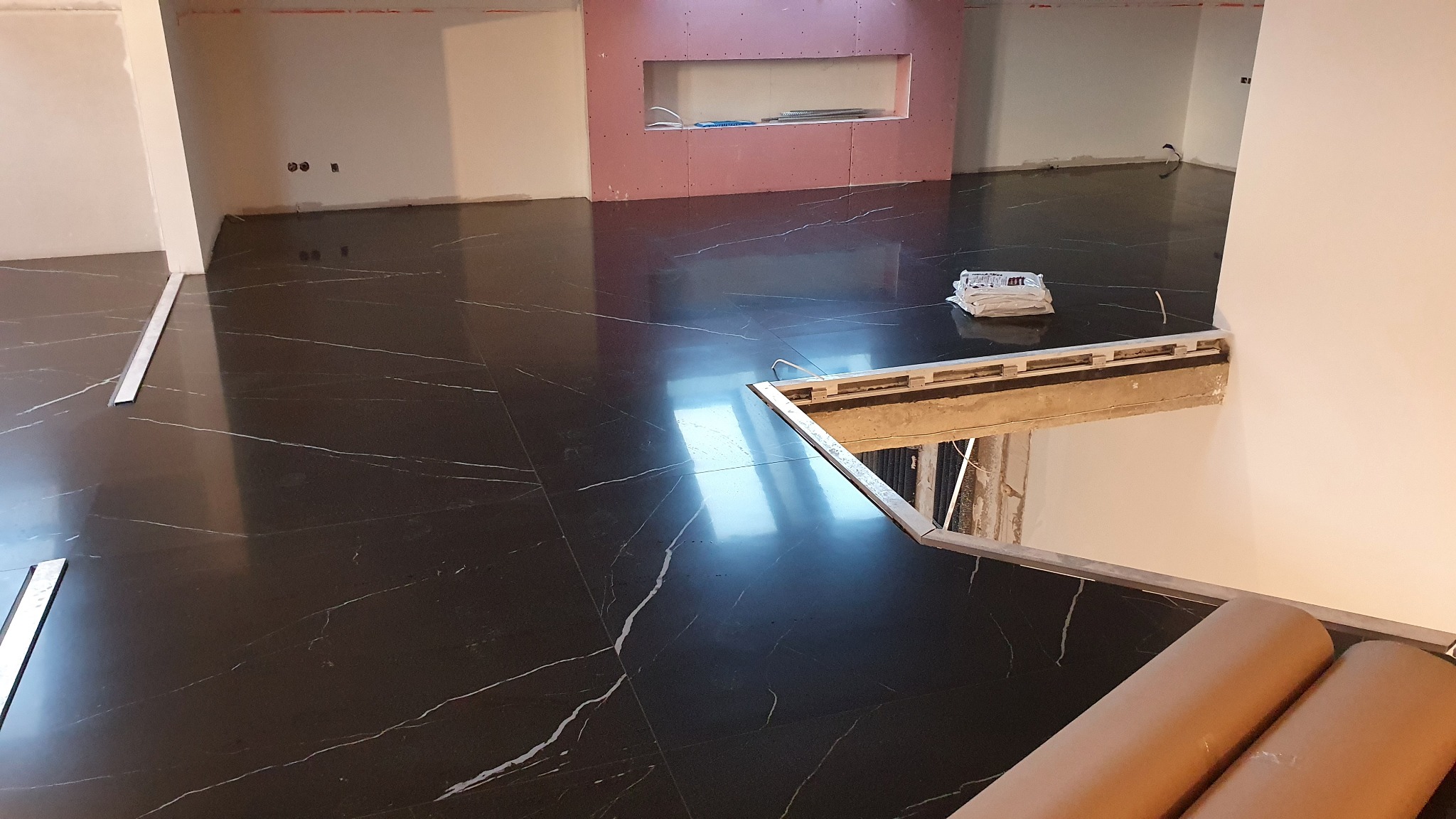 Położona, ciemna, marmurkowa posadzka w trakcie wykańczania, widoczne listwy przypodłogowe, niezabezpieczone schody i materiały budowlane w tle, ściany w trakcie przygotowania.
