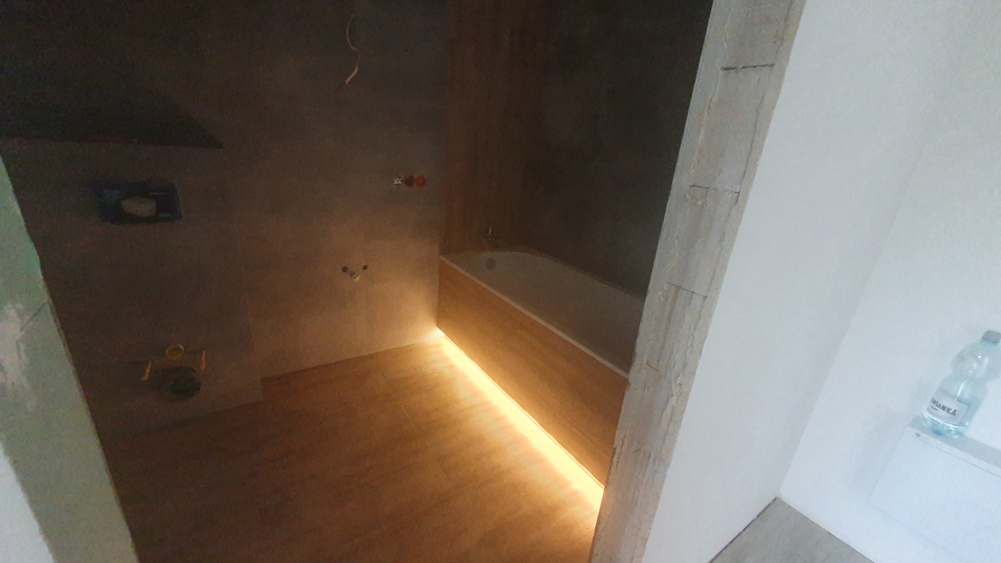 Surowa łazienka w trakcie remontu: widoczne niedokończone ściany, przygotowane instalacje, podświetlona wanna i drewnopodobna podłoga. Na ścianie butelka wody.