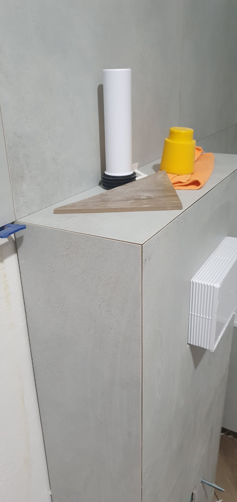 Szara, świeżo ułożona glazura na zabudowie w łazience, widoczne narzędzia glazurnicze: przyssawka, poziomica, dystanse, oraz biały syfon i żółty pojemnik.