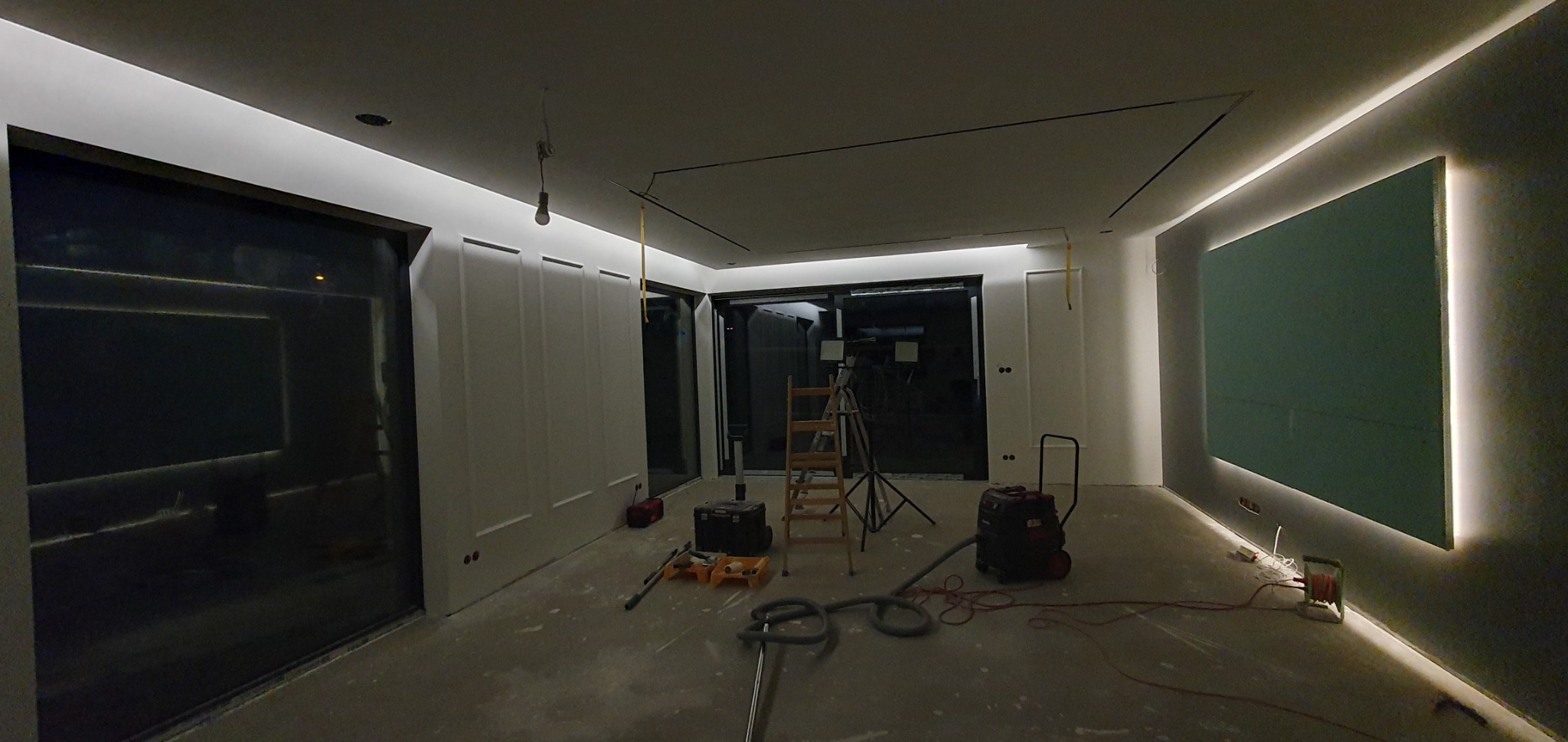 Wnętrze w trakcie wykańczania: widoczna płyta karton-gips z podświetleniem LED na ścianie, drabina, odkurzacz przemysłowy, narzędzia malarskie i surowe ściany z dekoracyjnymi panelami.