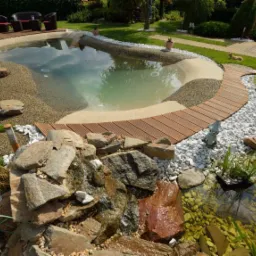 Oczko wodne z kaskadą kamienną, drewnianym pomostem i różnorodnym podsypanym kamieniem w ogrodzie. W tle widoczne meble ogrodowe.