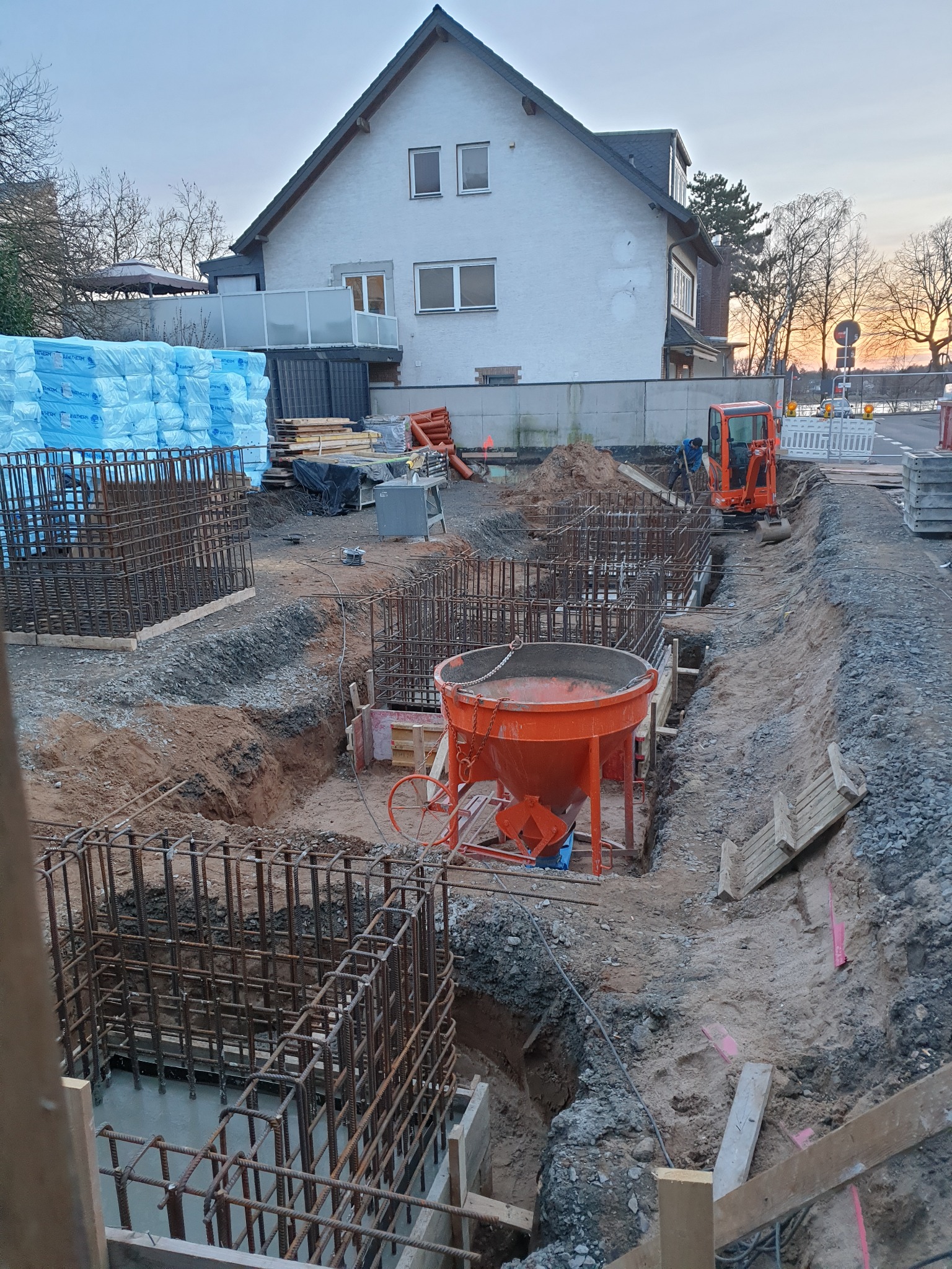 Wykop pod fundamenty nowego domu z widocznymi zbrojeniami, pomarańczową betoniarką i mini koparką w tle, na tle białego domu z balkonem i stertą niebieskiej izolacji termicznej.