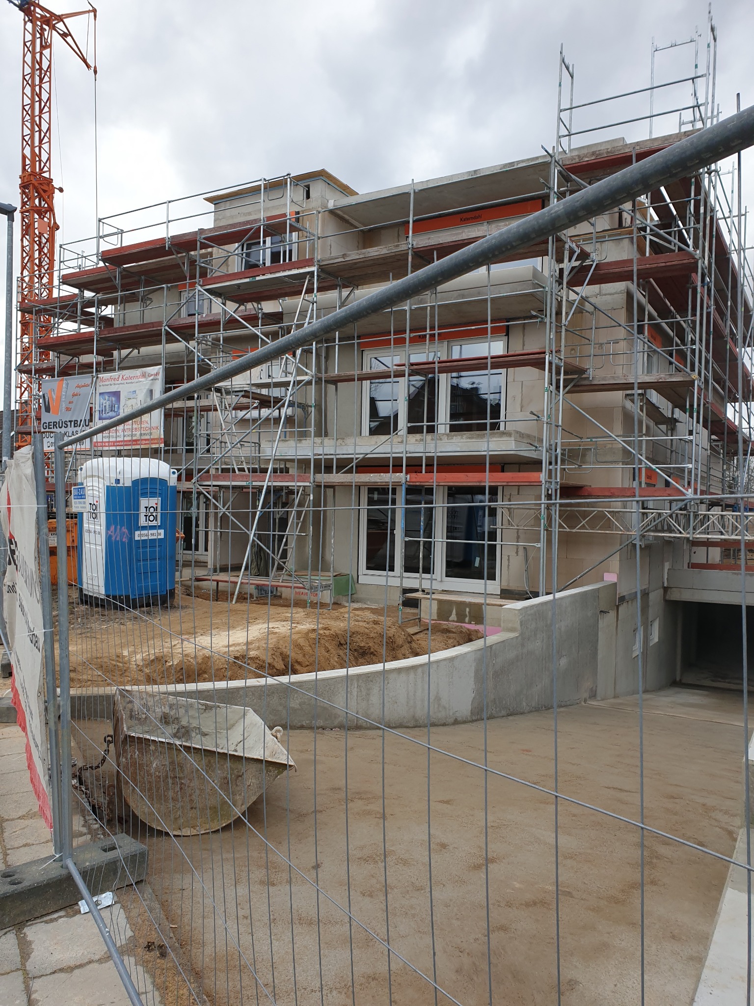 Surowy stan domu w trakcie budowy, z rusztowaniami, widoczny dźwig w tle, na pierwszym planie ogrodzenie budowy i kontener sanitarny Toi Toi.