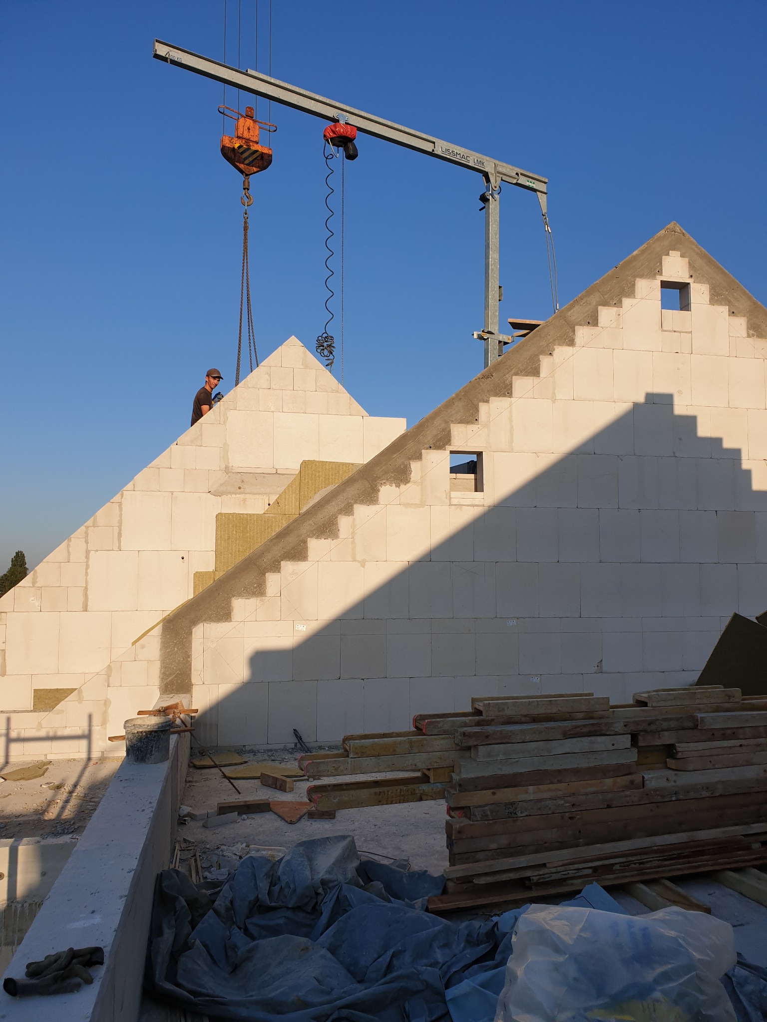 Budowa domu z białych bloczków, widoczny fragment ocieplenia, dźwig budowlany i pracownik na szczycie ściany, w tle błękitne niebo.