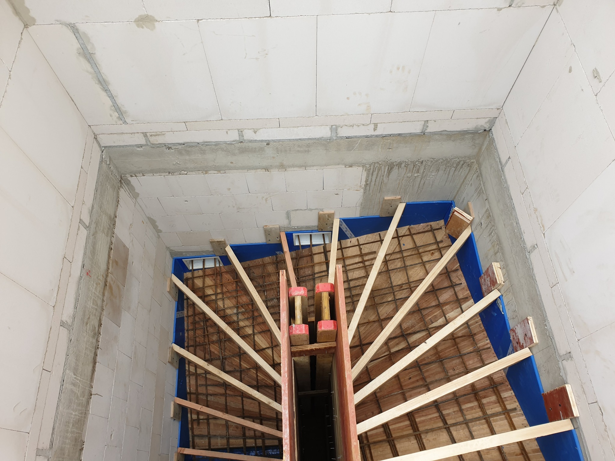 Wnętrze budynku w trakcie budowy, widok z góry na szalunek schodów z drewnianych belek i niebieskiej płyty, ściany z białych bloczków.
